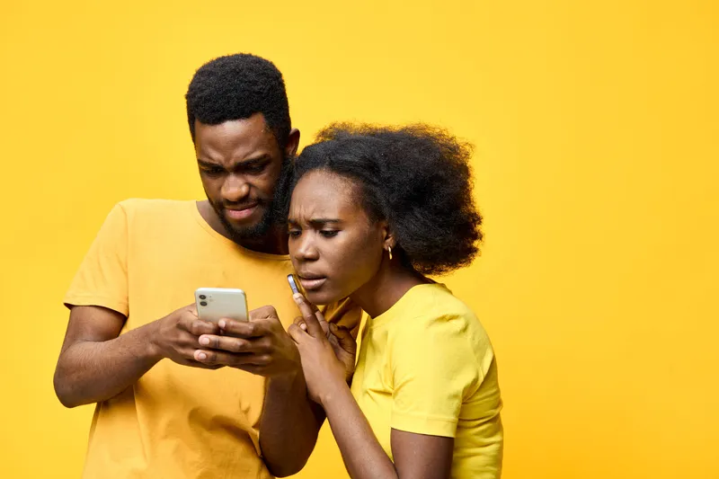 Concerned young couple checking their phone with worried expressions against a bright yellow background, showcasing emotions of anxiety and curiosity in modern fashion