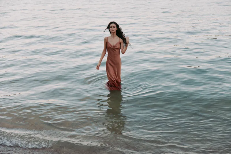 A woman stands in calm sea water near the shore, wearing a flowing rust dress as waves lap around her, capturing a serene moment of solitude and grace.