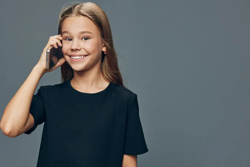 Girl smiling and talking on smartphone with hand near ear standing on gray background. Teenager with long hair in casual black shirt looking at camera.
