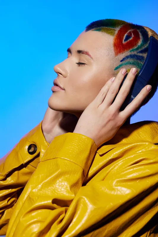 Woman with colorful artistic shaved hairstyle wearing yellow coat closes her eyes and touches head against blue background, showcasing unique fashion and self-expression.