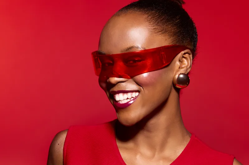 Smiling African American woman with dark skin wearing red futuristic sunglasses and red top against red background. Concept of fashion, style, beauty, and modern accessories.