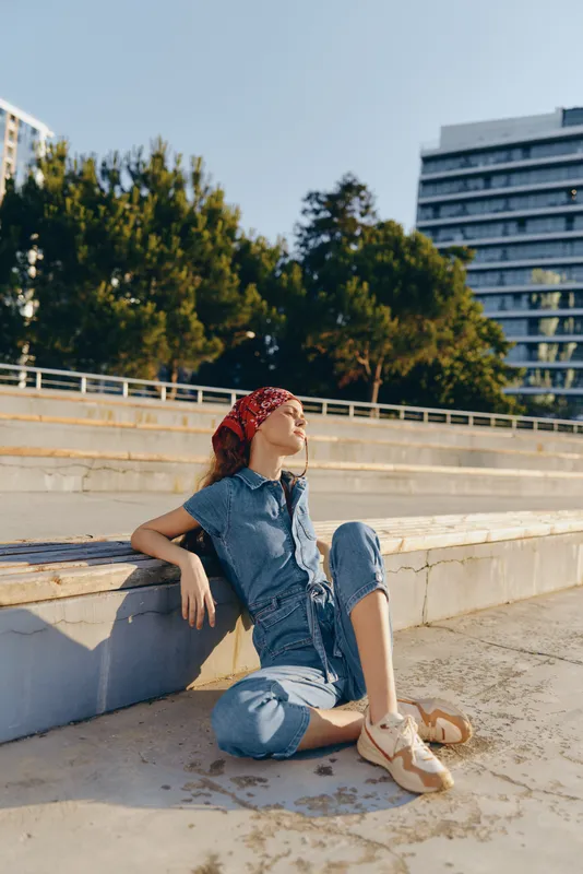 relaxed woman sitting outdoors on stone steps wearing casual denim jumpsuit and red headscarf enjoying sunny day with urban buildings and green trees in background
