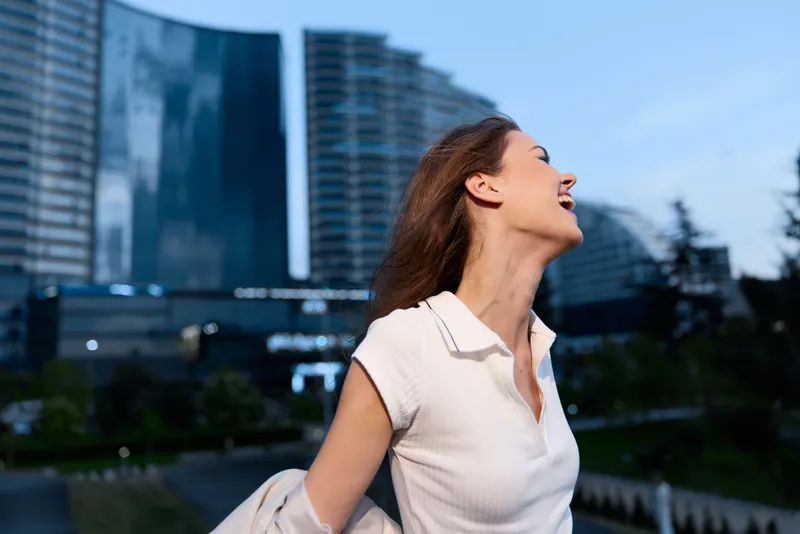 Joyful woman with long brown hair dressed in white shirt expressing happiness in urban cityscape with modern buildings and blue sky