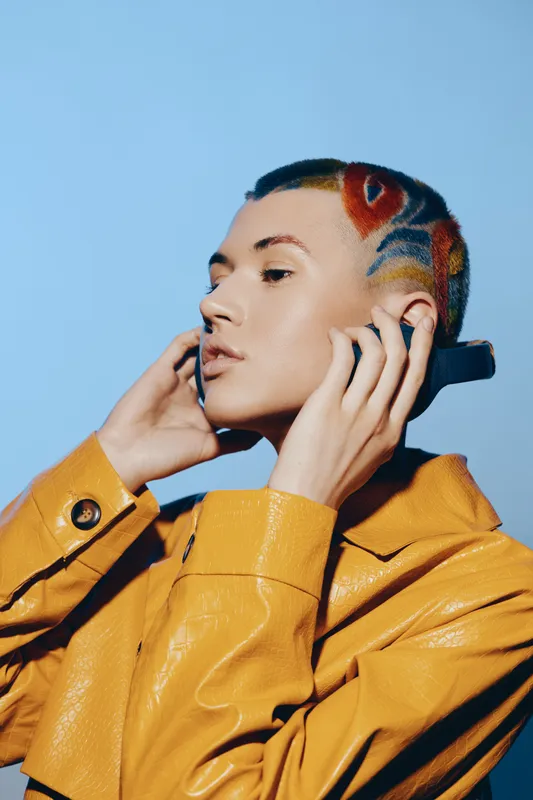 Young woman with colorful shaved hair in yellow jacket adjusting earbuds, showing modern style and confident expression against plain blue background.