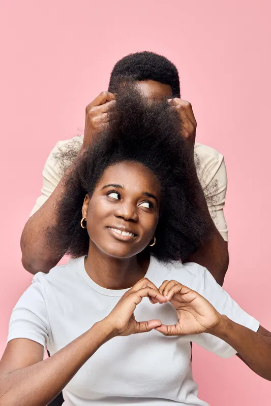 Happy couples expressing love with a heart gesture, showcasing natural hairstyles against a soft pink backdrop Afro hair, positive emotions, and playful interaction