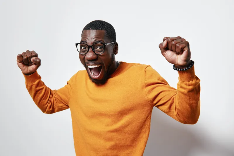 African American man with dark skin wearing glasses and orange sweater celebrating with raised fists and excited expression on white background in studio.