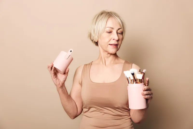 Mature woman with short grey hair holds makeup brushes and container in beige top against light background. Concept for beauty routine, self-care, cosmetics, and natural skincare products.