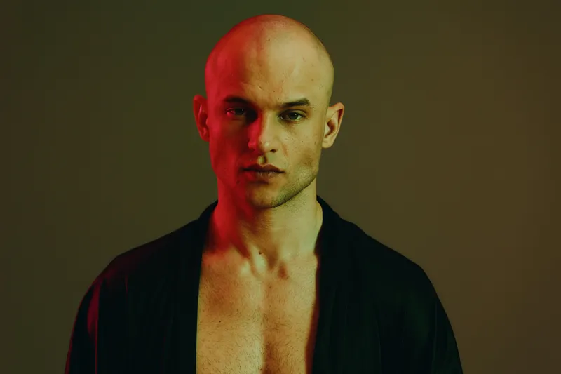 Man in a dark robe with striking lighting, showcasing intense facial expression and unique color contrasts. A compelling representation of modern masculinity.