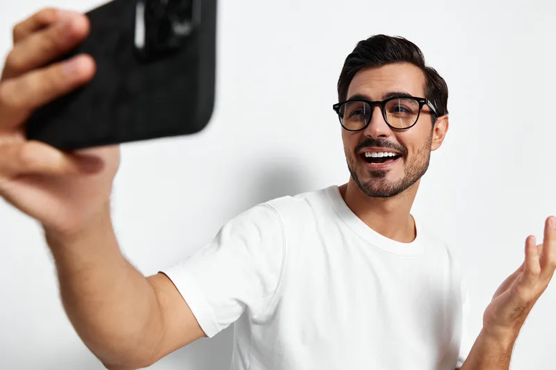 Smiling man wearing glasses takes a selfie with his mobile phone while sitting against a plain white background. Vibrant lifestyle moment captured in studio portrait.