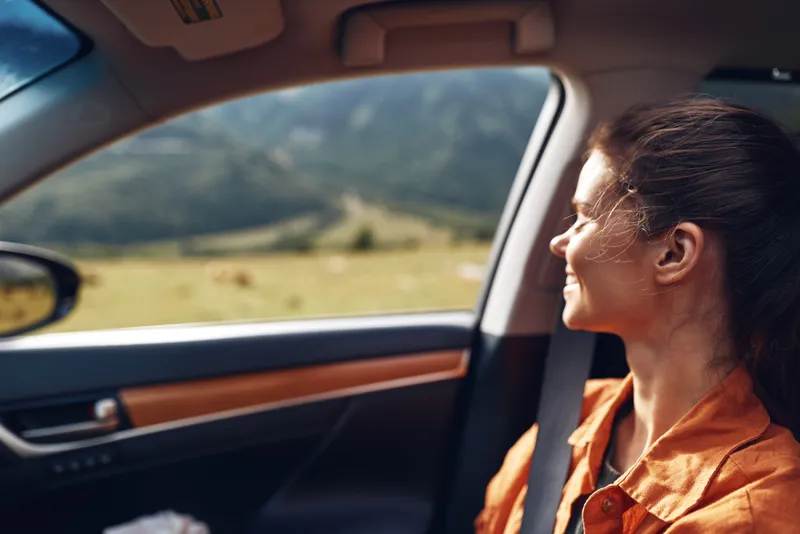 Woman in an orange shirt sits as a passenger in a car, gazing through the window at a vast scenic landscape during a calm travel moment.