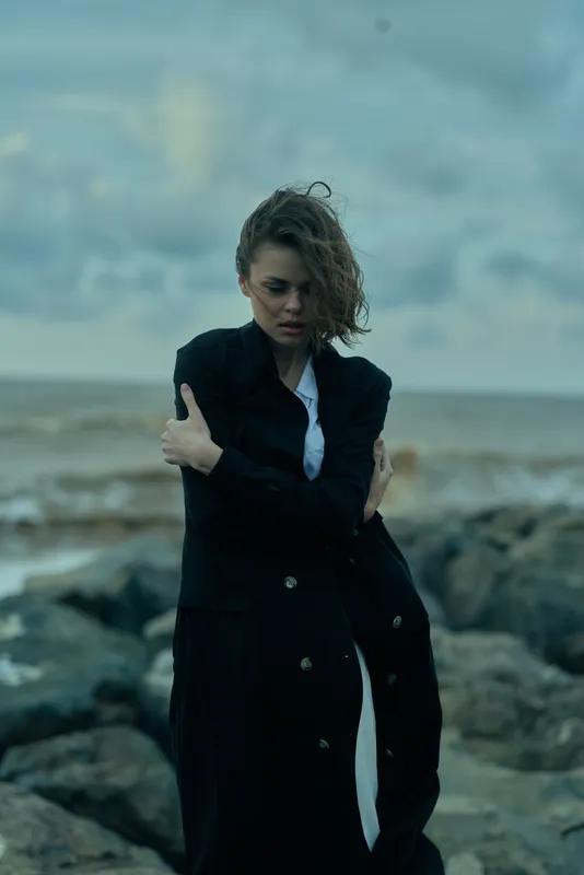 Woman in a long dark coat on a rocky shoreline, windblown hair, moody seascape highlighting fashion and resilience, dramatic palette.
