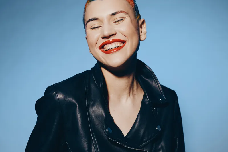 smiling woman with red lipstick wearing black leather jacket against blue background, expressing joy and confidence, vibrant portrait with bright colors