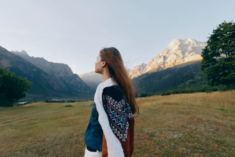 Woman in a meadow looks toward mountains in a wide landscape, wearing a patterned shawl; travel portrait of nature, solitude and serene outdoor exploration in golden light.