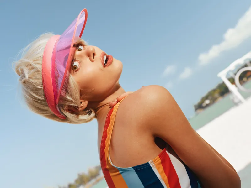 A stylish beach portrait featuring a sunlit woman in a striped swimsuit and vivid visor, captured from a low angle against a clear blue sky and bright sandy backdrop.