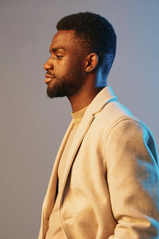 Stylish African man in profile wearing a beige blazer, against a soft blue grey background, conveying confidence and modern fashion for editorial use
