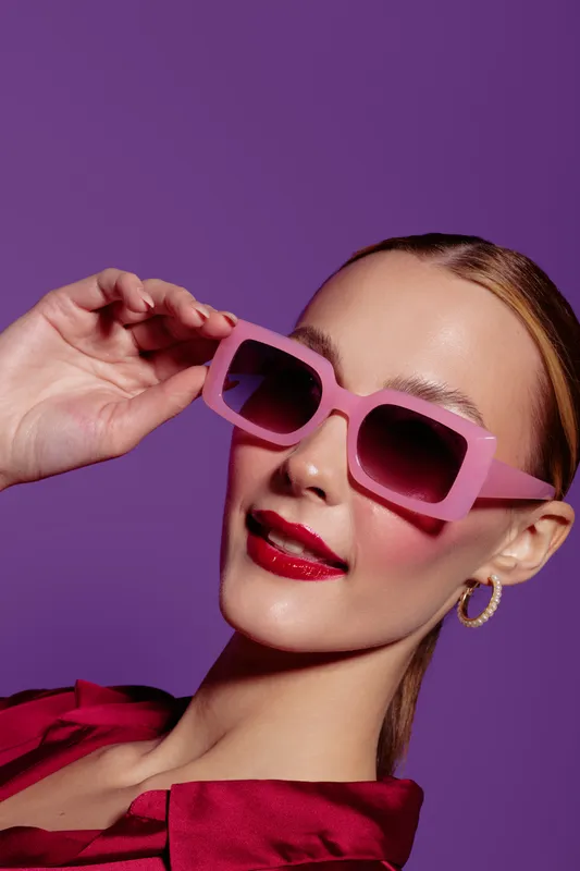 Stylish woman with pink sunglasses and red lipstick poses against purple background. Fashion portrait with trendy accessories and confident expression. Concept of glamour and modern style.