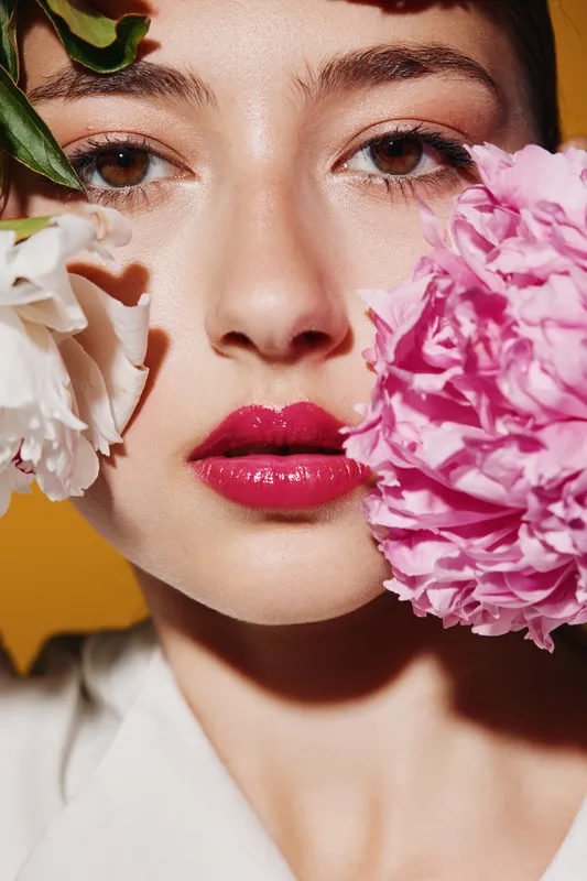 Close-up beauty portrait of young woman with glossy pink lips and natural makeup holding white and pink flowers near face on vibrant orange background. Creative fashion concept.