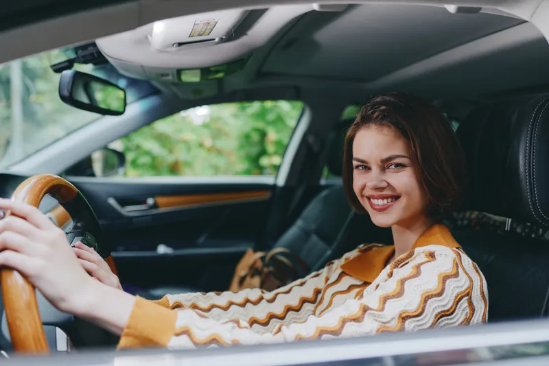Smiling woman driver in a modern car, seated behind the wheel with a cheerful expression. Warm lighting highlights the interior and steering area.