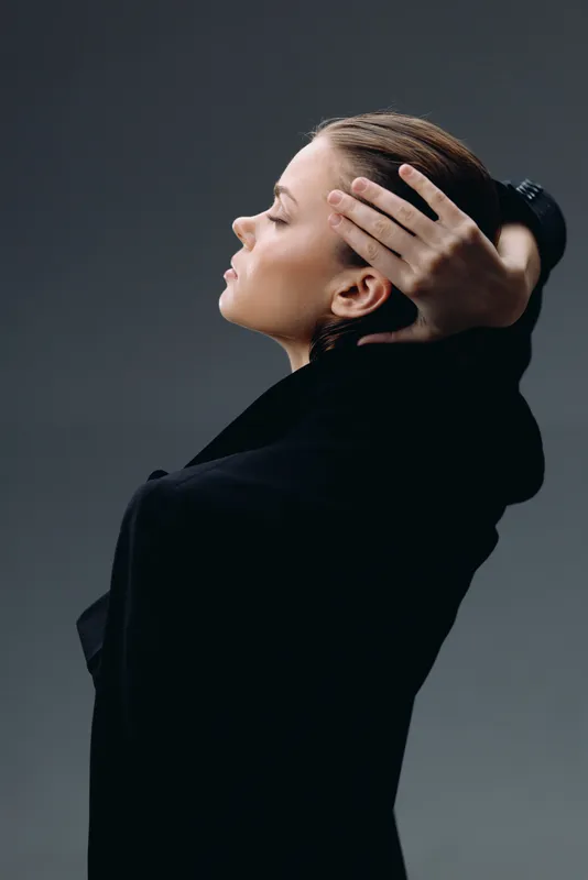 Profile of a thoughtful woman in black clothing with closed eyes and hand in hair, portrait with soft lighting and gray background.