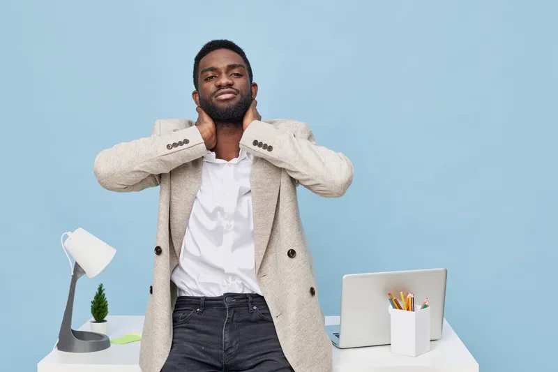 Confident happy African American man standing at modern workspace with hands near neck, smiling, wearing beige blazer and white shirt, isolated on light blue background, studio portrait. People lifest