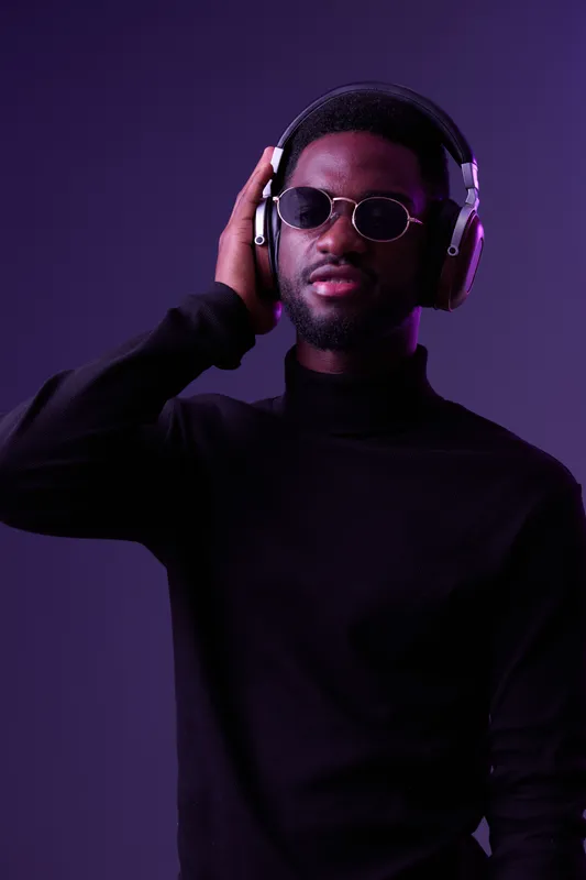 Stylish young Black man wearing headphones and sunglasses, enjoying music against a vibrant purple background