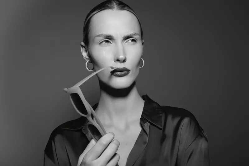 Portrait of a serious white woman with light skin holding sunglasses and looking away. Black and white studio image with dramatic lighting.