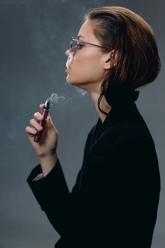 Young woman vaping electronic cigarette with gray background in dark outfit, contemplative mood, modern lifestyle concept.