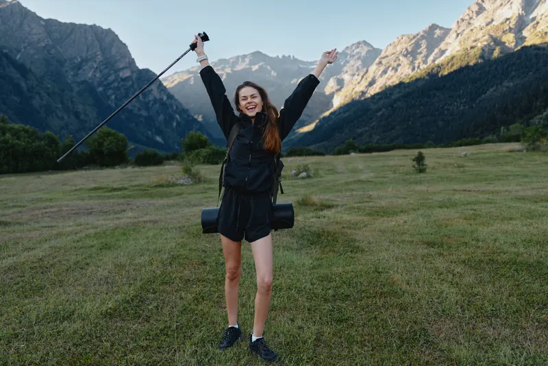 A cheerful hiker stands in a grassy valley with towering mountains in the background, raising a trekking pole and smiling, enjoying an adventurous outdoor moment.