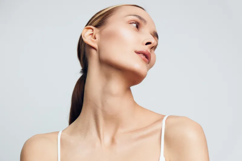 Young woman portrait with serene expression, looking upward, exposed neck and clean skin, wearing white top, isolated on light gray background, soft lighting enhances natural features