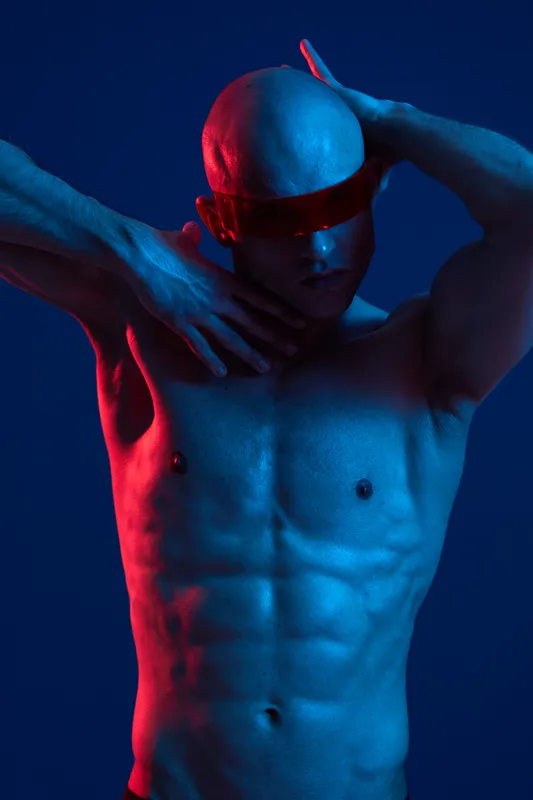Athletic man posing dramatically in vibrant blue and red lighting, showcasing a strong physique and wearing a stylish visor. Captivating blend of color and expression.