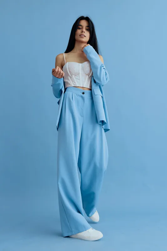 Stylish woman wearing light blue suit and white lace top standing confidently against a plain blue background with casual white sneakers, showcasing modern fashion.