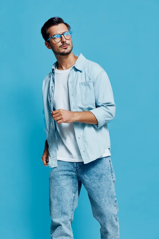 Stylish young man wearing blue shirt and glasses, standing confidently against a vibrant blue backdrop, displaying a trendy casual look. Fashion lifestyle concept
