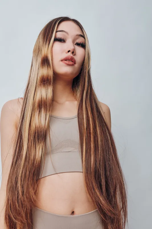 Portrait of young woman with long straight hair and neutral expression wearing beige sleeveless top standing against plain background in studio lighting.