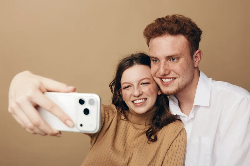 Smiling young couple takes selfie with smartphone on beige background. Happy man and woman enjoy moment together, close pose, casual clothing, natural expressions, modern lifestyle concept.
