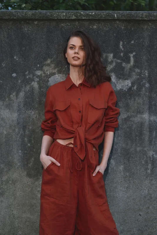 Vibrant woman outdoors wearing rust shirt and matching trousers, relaxed pose with hands in pockets, natural light, urban backdrop, casual lifestyle moment with soft shadows