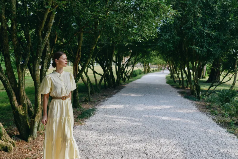 A woman in a flowing yellow dress walks along a gravel path bordered by trees, enjoying a peaceful outdoor park scene with dappled sunlight.