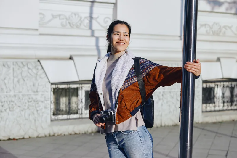 Woman with camera smiles beside a street pole, wearing a knit scarf, colorful jacket, and a backpack, captured in bright daylight for street fashion and outdoor photography.