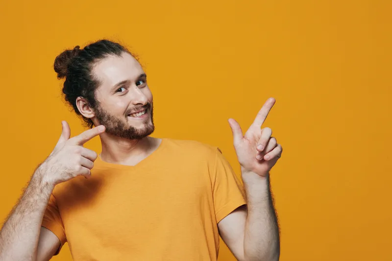 Cheerful young man in casual orange t shirt pointing with both hands on bright yellow background, showcasing happiness and positivity