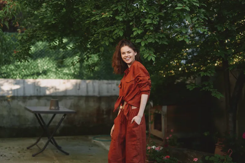 A smiling woman in a red dress stands in a sunlit garden, conveying a casual lifestyle moment outdoors, with leafy backdrop, relaxed pose, and warm daylight