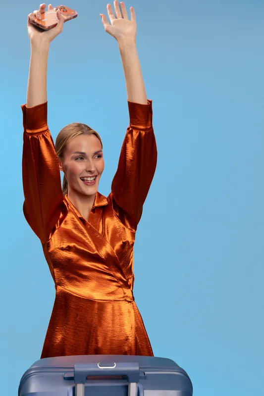 Smiling woman in orange satin dress raises hands in excitement behind blue suitcase. Blue background highlights joyful travel concept and vacation preparation with happy female traveler.