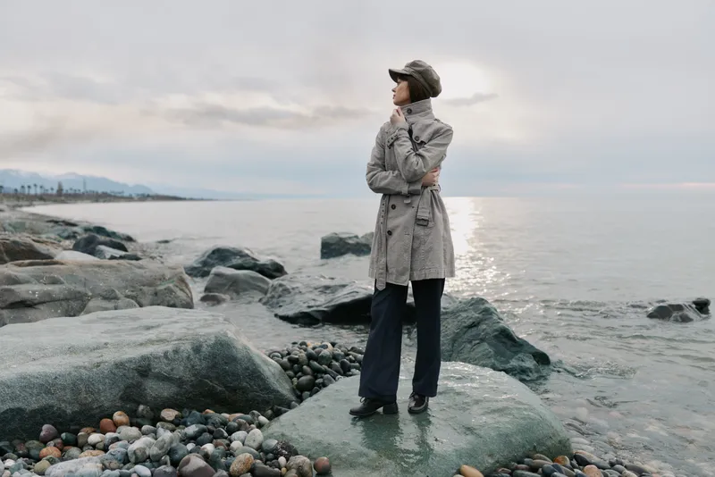 Woman in grey coat and hat stands on rocky beach near calm sea, looking into distance. Overcast sky and peaceful atmosphere create mood of contemplation and solitude by water.
