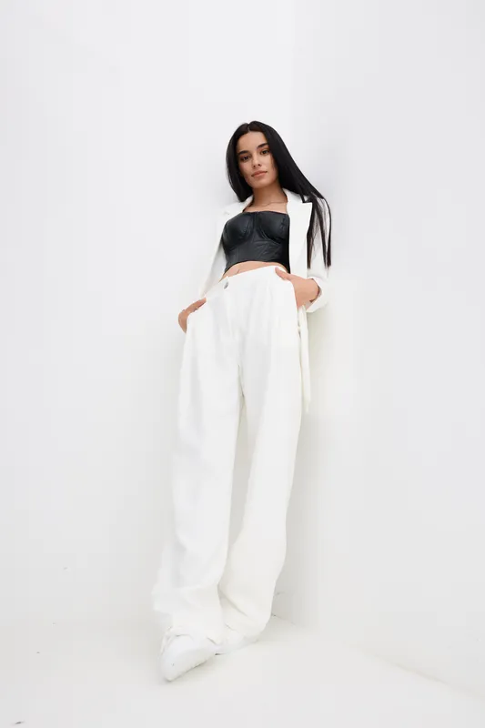 tall woman in white pants and black top stands against a white wall with hands in pockets, showcasing modern stylish casual outfit and relaxed confident pose indoors