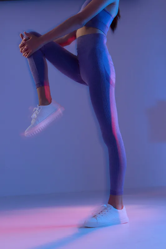 Woman sport fitness in purple textured leggings stretching leg, white sneakers, preparing for workout in moody blue and pink light, active lifestyle simplicity.