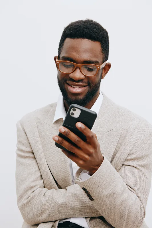 Smiling man with glasses using smartphone, casual beige blazer over white shirt, joyful expression, modern technology, light background, portrait, lifestyle concept