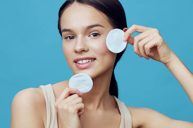 Young woman holding cotton pads, promoting skincare and healthy beauty routine, smiling on blue background, natural and fresh appearance