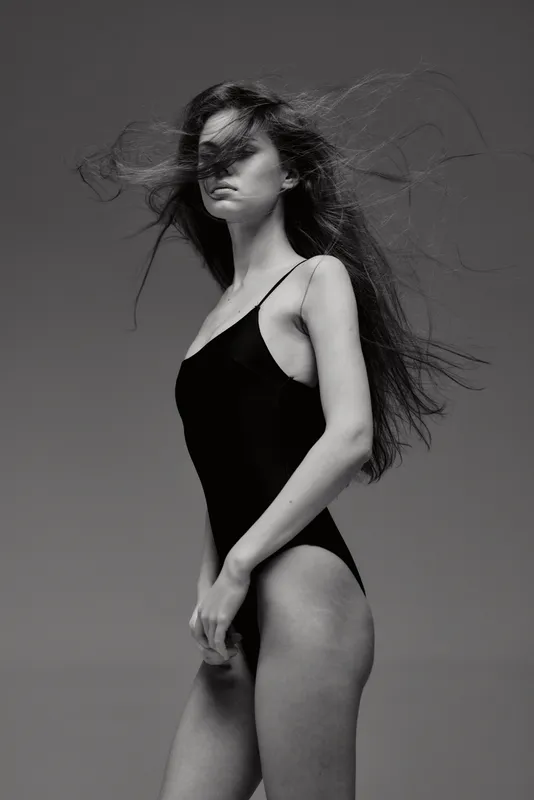 Black and white portrait of a woman in a black bodysuit with windblown hair, expressing elegance and strength.