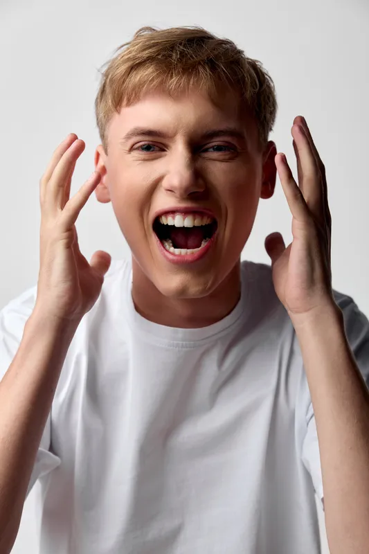 Energetic young man shouting with open mouth and raised hands, expression of excitement and surprise, casual white tee, studio lighting, dynamic emotion