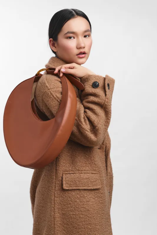 Asian woman stands in a clean studio, wearing a tan wool coat and carrying a round brown bag, exuding chic confidence, poised elegance, and a timeless fashion vibe.