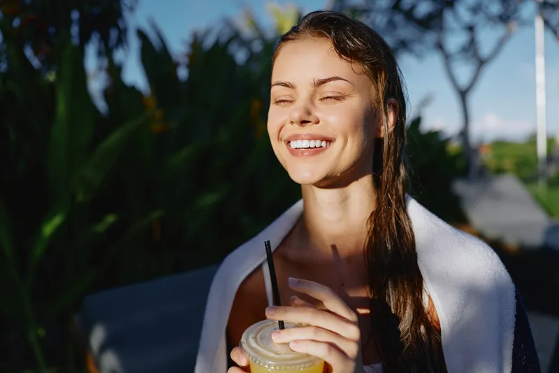 A smiling woman enjoys a cold beverage outdoors on a sunny day, with a towel draped over her shoulders. A relaxed lifestyle moment in natural light.