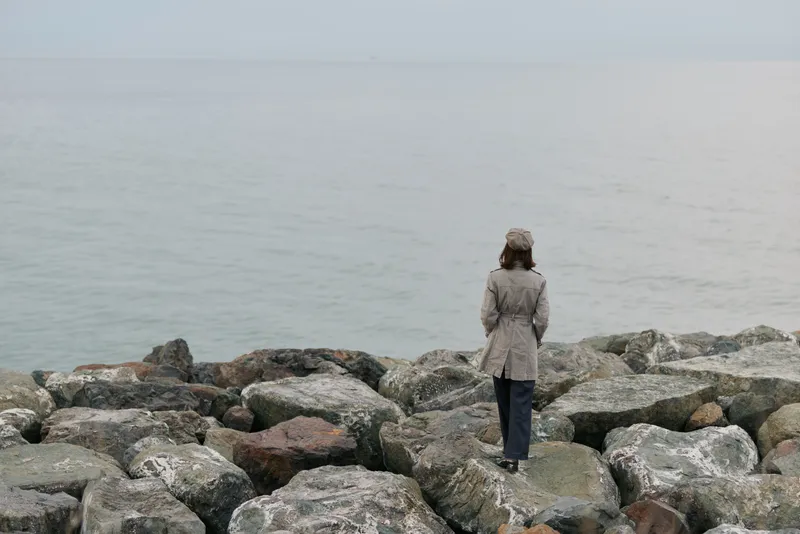 A woman in a coat and hat walks alone on rocky shore facing calm sea. Overcast sky sets a quiet, reflective mood with natural, muted colors and peaceful coastal atmosphere.